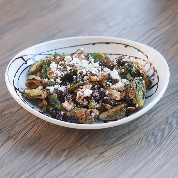 A white bowl of roasted Brussels sprouts with feta cheese and balsamic glaze on a wooden table.