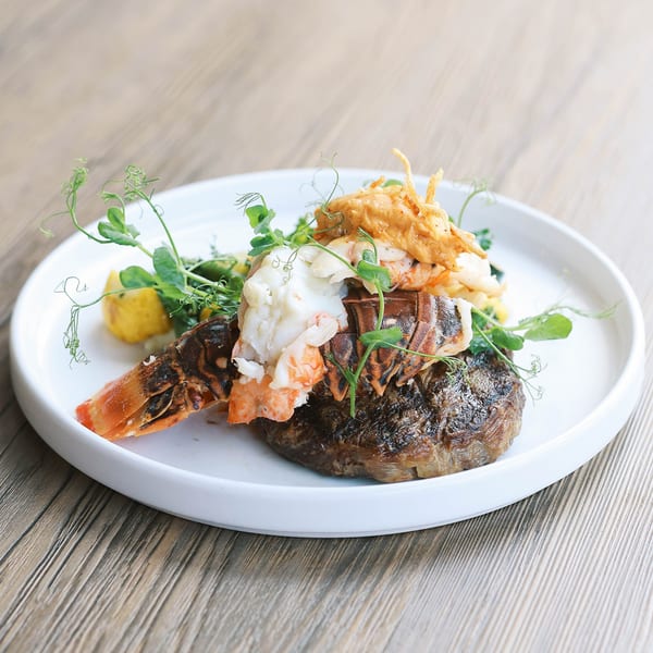 A grilled steak topped with lobster tail, pea shoots, and a creamy sauce on a white plate.