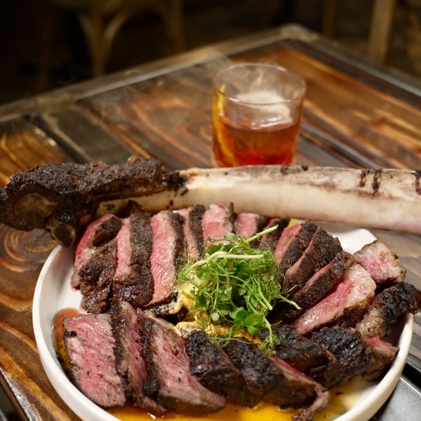 Sliced tomahawk steak with microgreens and a drink on a rustic wooden table at The Rustik Fork.
